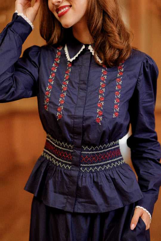 A woman wearing the Ivy City Co Bluebell Top in Sapphire Rosette Embroidery, featuring a white collar and a patterned waistband, smiles as she touches her hair. The softly blurred background highlights the blouse’s delicate smocked waist details.