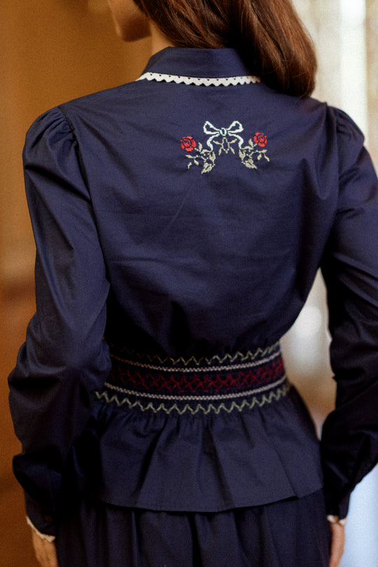 A girl is wearing the Ivy City Co Bluebell Top in Sapphire Rosette Embroidery, a navy blue holiday blouse featuring a wide, smocked waist and a bow detail on the back. The photo is taken from behind.