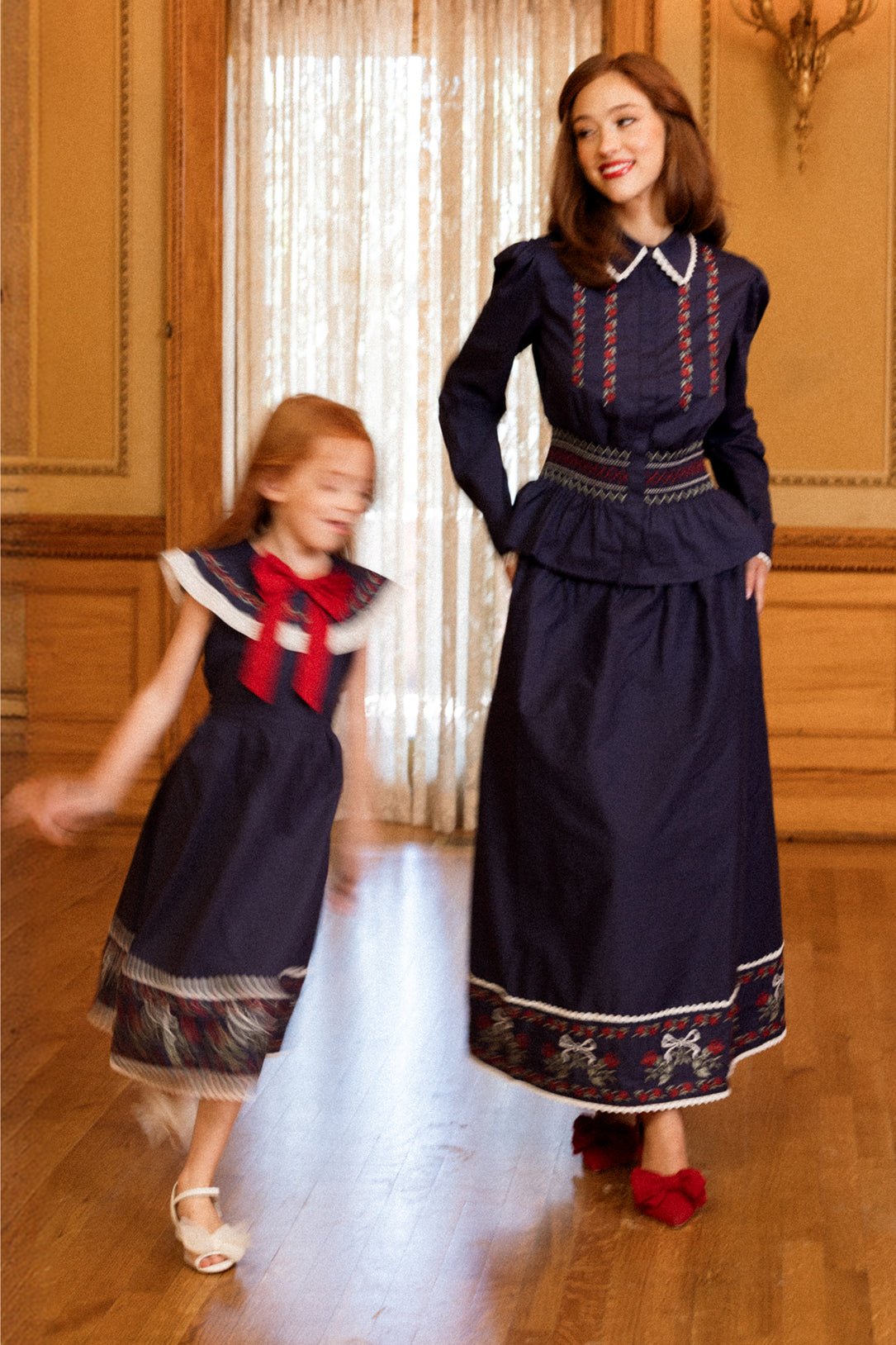 A woman and a young girl in matching navy dresses with red and white trim stand in an ornate room with wooden floors. The girl, wearing the Ivy City Co Bluebell Top in Sapphire Rosette Embroidery, is spinning, creating a sense of motion, while the woman smiles and looks at her.