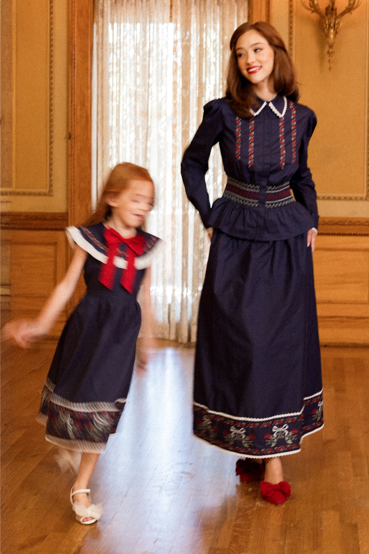 A woman and a young girl in matching navy dresses with red and white trim stand in an ornate room with wooden floors. The girl, wearing the Ivy City Co Bluebell Top in Sapphire Rosette Embroidery, is spinning, creating a sense of motion, while the woman smiles and looks at her.