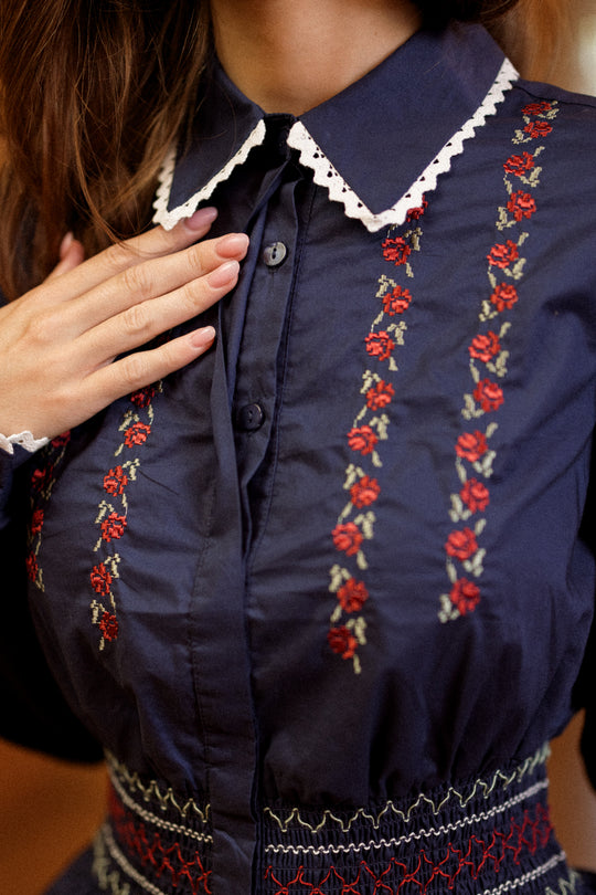 An individual wearing the Ivy City Co Bluebell Top in Sapphire Rosette Embroidery—featuring a navy blue smocked waist, white lace-trimmed collar, and delicate sapphire rosette embroidery—places their hand on their chest.