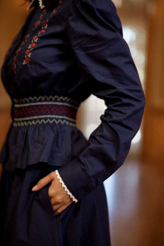 A person wears the Ivy City Co Bluebell Top in Sapphire Rosette Embroidery, featuring a patterned waistband. One hand is tucked in the pocket, with a softly blurred background.
