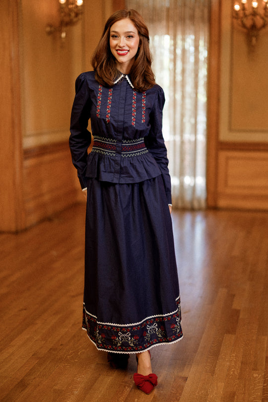 A woman stands indoors on a wooden floor, smiling with her hands at her sides, wearing the Bluebell Top in Sapphire Rosette Embroidery by Ivy City Co paired with red shoes adorned with bows.