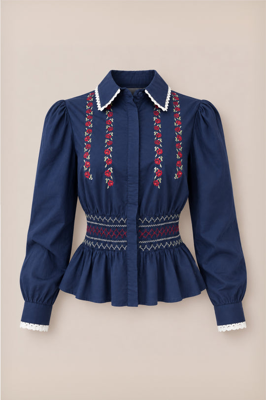 Introducing the Bluebell Top in Sapphire Rosette Embroidery by Ivy City Co—a navy blue girls’ holiday blouse featuring long sleeves with puffed shoulders, elegant white lace trim, and delicate sapphire rosette embroidery on the front. This cotton top showcases a smocked fitted waist, a peplum hem for added charm, a collared neckline, and completed with button closures.