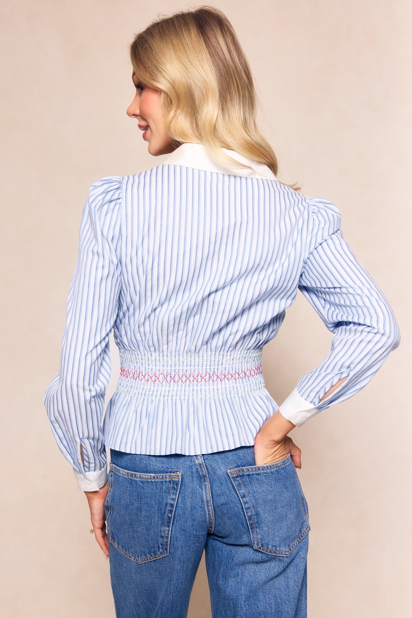 A woman with loose blonde hair faces away, wearing the Ivy City Co Bluebell Top in Blue & White Stripes with white cuffs and collar, paired with blue jeans. The nursing-friendly blouse allows easy access; her right hand is on her hip.