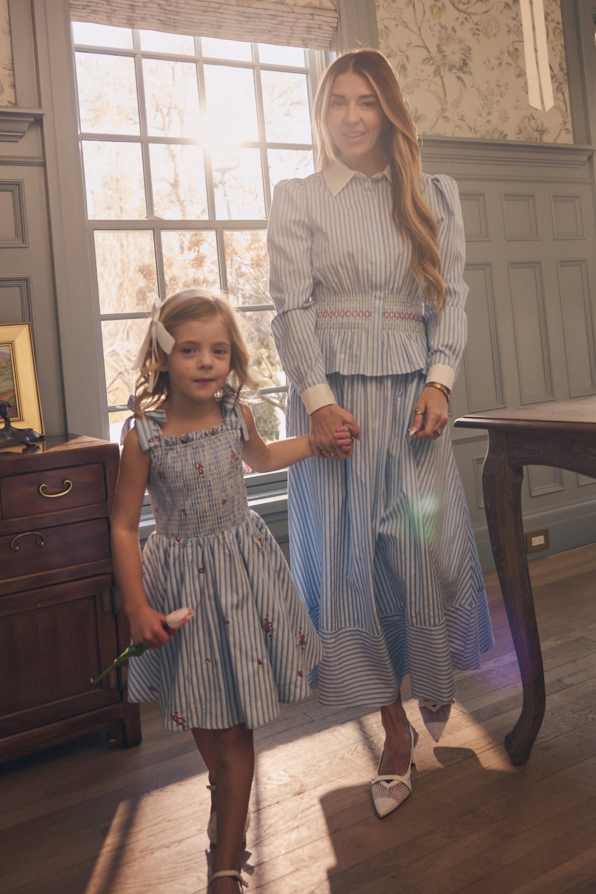 A woman and young girl stand indoors holding hands, both wearing Ivy City Co’s Bluebell Skirt in Blue & White Stripes. Sunlight pours through a large window behind them, creating a warm, inviting scene.
