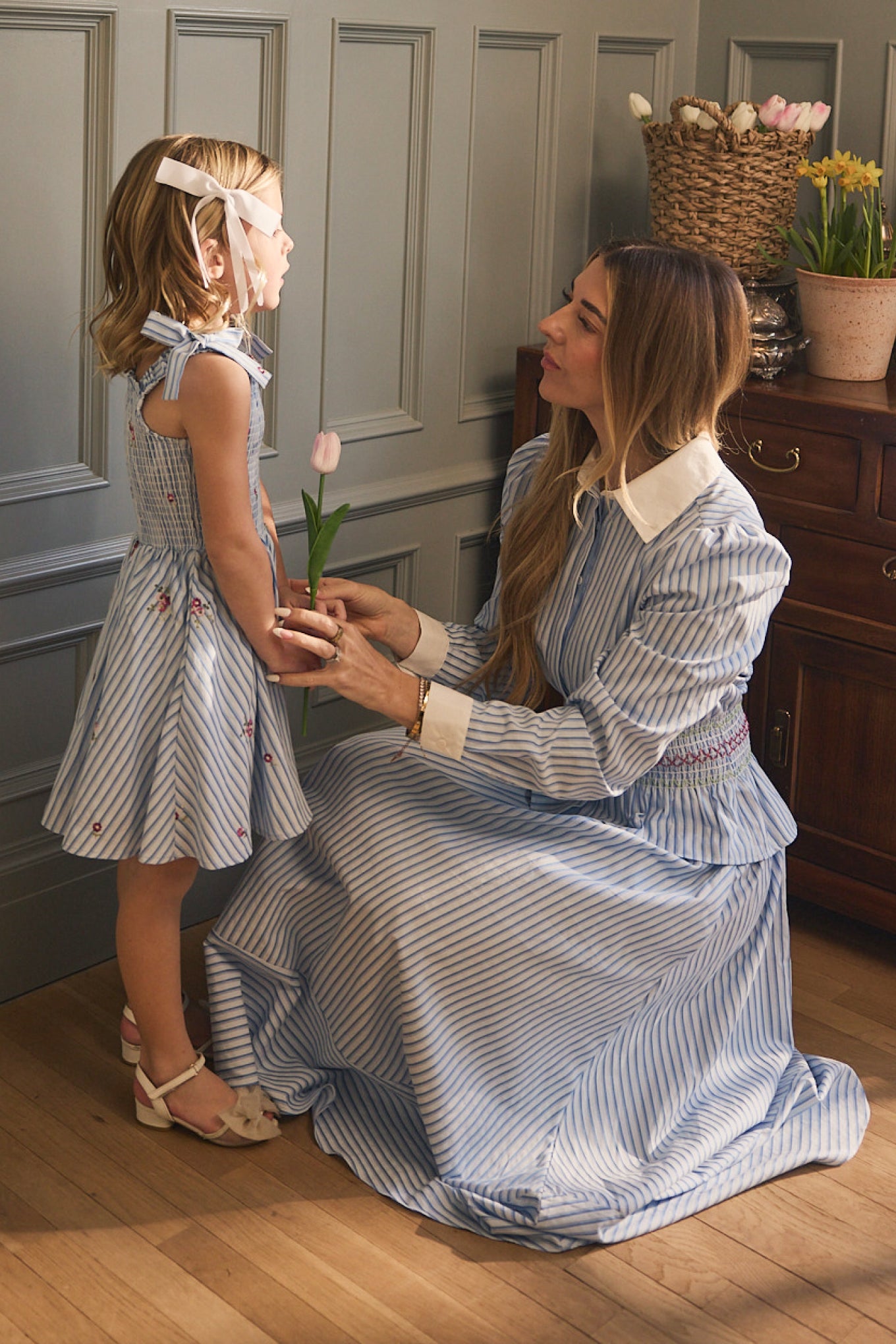 A woman and a young girl smile together; both wear Ivy City Co's Bluebell Top in blue & white stripes, featuring white collars, in a gray-paneled room with flowers on a wooden cabinet. The girl holds a tulip.