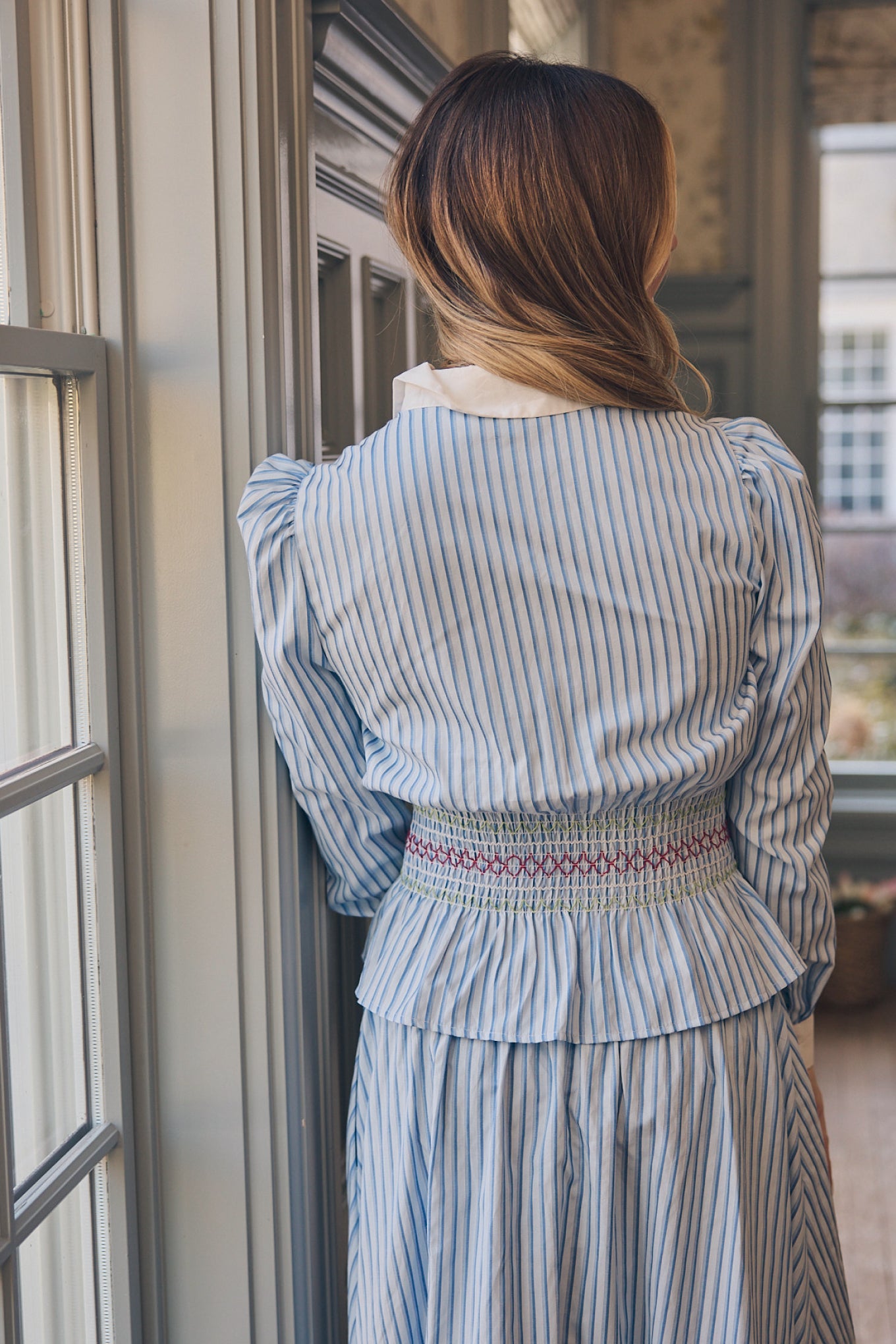 A woman with light brown hair stands indoors by a window and painted wooden wall, facing away while wearing the Ivy City Co Bluebell Top in blue & white stripes.