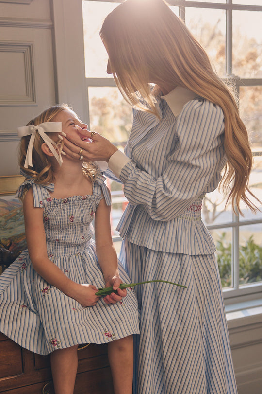 A woman and a young girl smile at each other in a sunlit room, both wearing Ivy City Co's Bluebell Top in blue and white stripes, featuring smocked waistbands. The woman gently touches the girl's face as she holds a green stem.