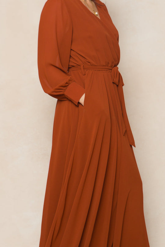 A woman models the Andie Dress in Amber Chiffon by Ivy City Co, featuring long sleeves, a soft and flowy rust-colored chiffon fabric, and a matching belt tied at the waist. With one hand in the pocket and set against a light, neutral background, this women’s maxi dress blends comfort and elegance—ideal for fall bridesmaids or as an elevated everyday look.