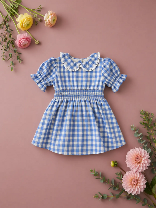 The Baby Hannah Dress Set in Blue Gingham by Ivy City Co features puffy sleeves, a hand-smocked bodice, and a lace-trimmed collar. It’s displayed on a pink surface with yellow, pink, and peach flowers amid green foliage.