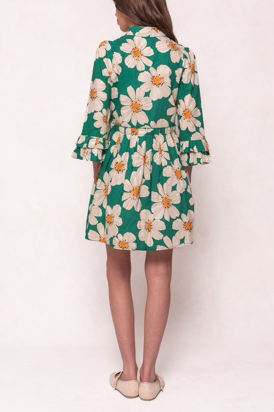 A person faces away wearing Ivy City Co’s Bowie Dress in Green with White Floral, featuring ruffled sleeves and beige flats; the cotton, knee-length dress stands out softly against a light, plain background.