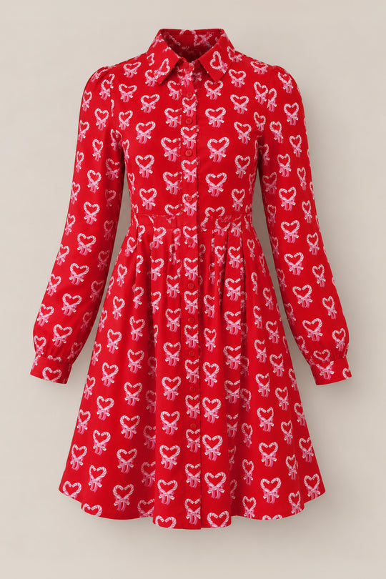 Introducing the Bowie Long Sleeve Dress in Poinsettia Candy Canes by Ivy City Co. This cotton holiday dress features a red base with an all-over print of pink and white candy canes shaped into hearts. Designed with long sleeves, a classic collar, and a button-down front for nursing-friendly wear, it also boasts a slightly pleated skirt. The dress is presented against a plain, light background.