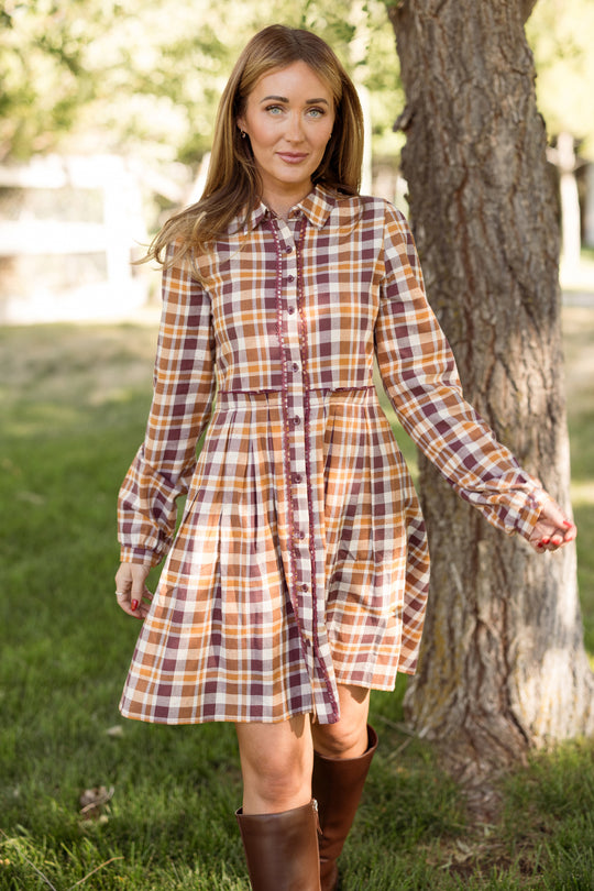 A woman stands outside near a tree, gently holding her Ivy City Co Bowie Long Sleeve Dress in Spiced Plaid and smiling softly. She pairs the dress with brown boots, surrounded by grassy scenery, trees, and warm sunlight.
