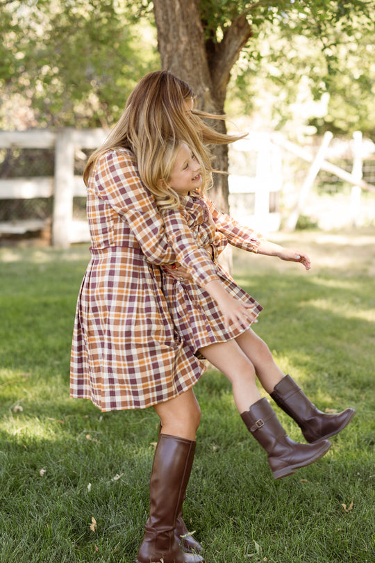 A woman spins a smiling young girl in her arms outdoors. Both wear matching Ivy City Co Bowie Long Sleeve Dresses in Spiced Plaid with brown boots, standing on green grass with trees and a fence in the background.