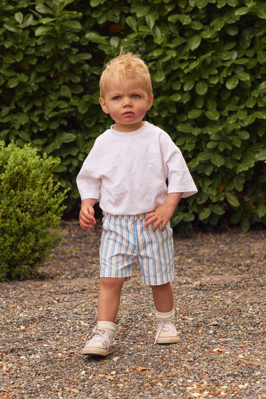 A young child with blonde hair stands outdoors on a gravel path, wearing a loose white T-shirt and Ivy City Co’s Boys’ James Shorts in Heritage Stripe with an elastic waistband. Socks, sneakers, and green bushes complete the scene.