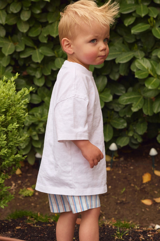 A young blond child wears a white oversized T-shirt and Ivy City Co Boys' James Shorts in Heritage Stripe, featuring an elastic waistband, while standing outside near green bushes with a thoughtful expression.