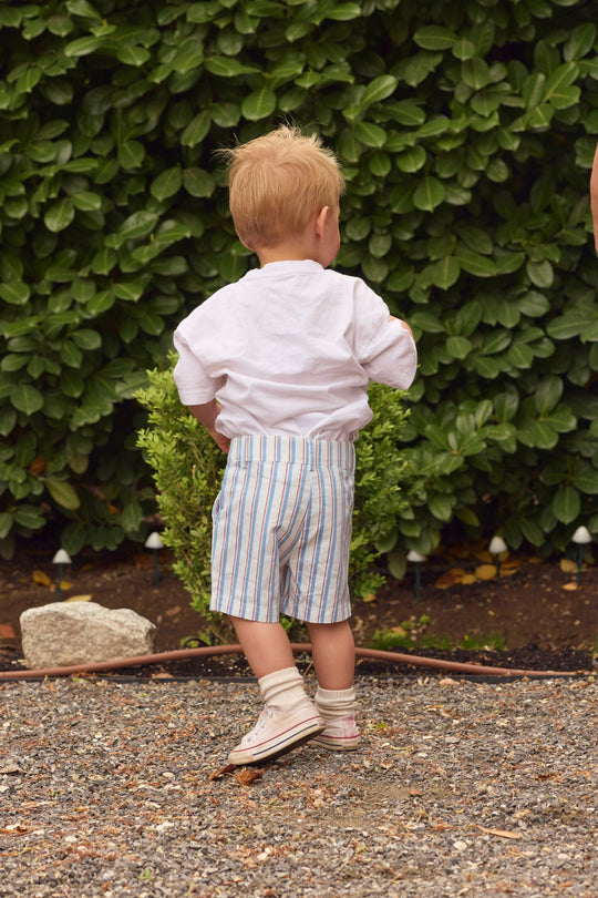 A young child with short blond hair and a white shirt wears Ivy City Co's Boys' James Shorts in Heritage Stripe, featuring an elastic waistband, standing on gravel with green leafy bushes in the background, facing away from the camera.