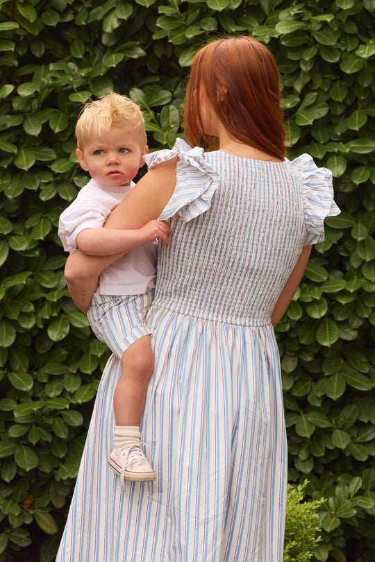 A woman with long red hair holds a blond toddler as they stand by a leafy green hedge, both wearing matching Ivy City Co Boys' James Shorts in Heritage Stripe with elastic waistbands.