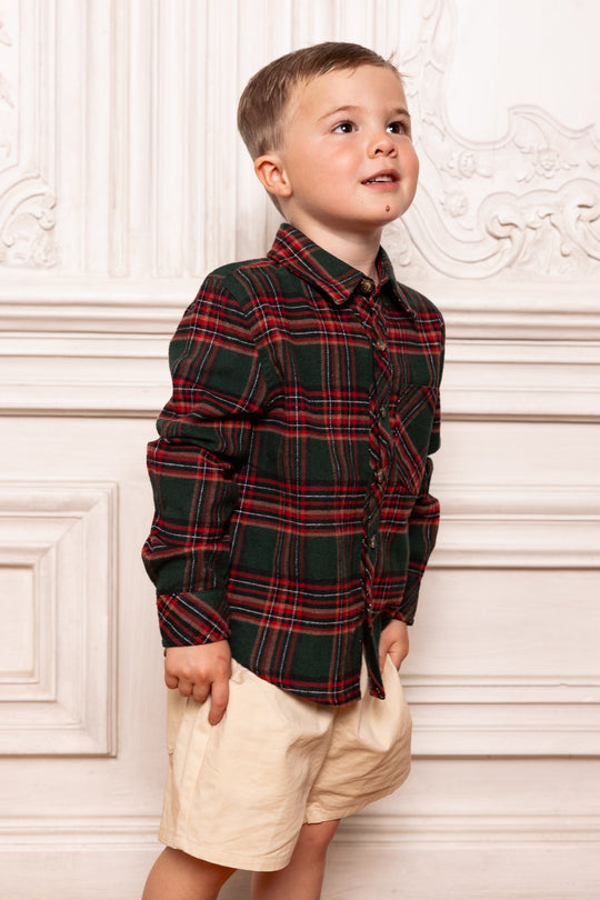 Child wearing a plaid shirt and shorts standing against a decorative white wall.
