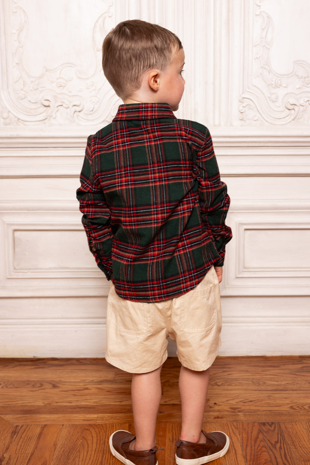 Child wearing a plaid shirt and beige shorts standing against a decorative wall.