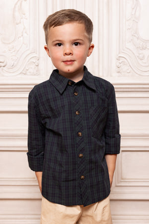 Young boy wearing a dark plaid shirt with buttons against a decorative white wall.