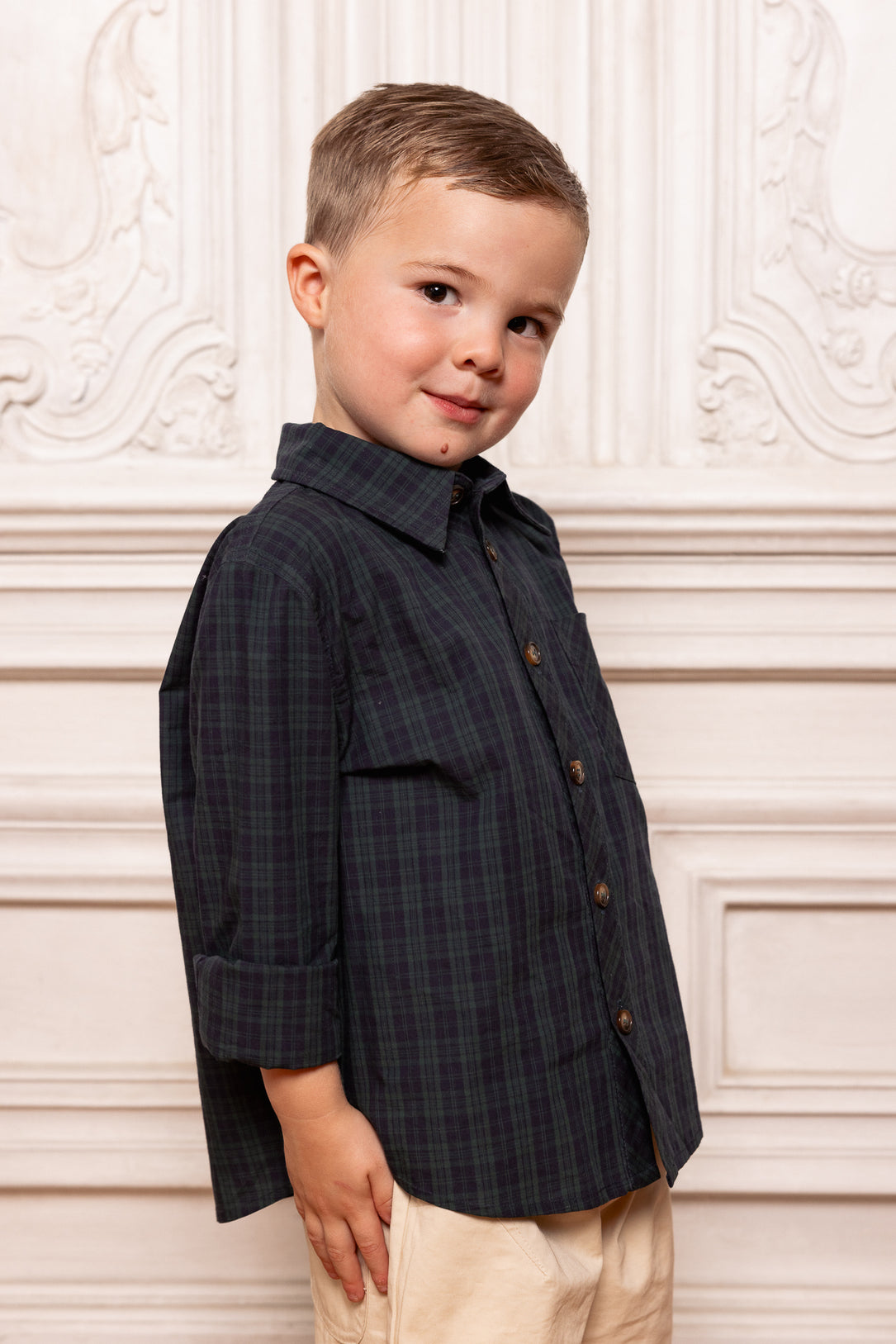 Young boy wearing a dark plaid shirt against a decorative white wall.