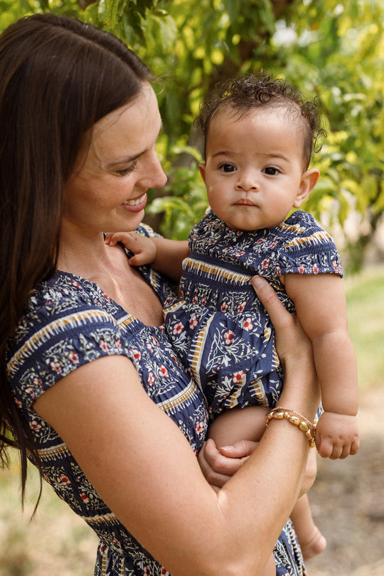 A woman smiles outdoors while holding a baby dressed in the Ivy City Co Baby Marlow Romper in Navy Fleur. Both wear matching blue, patterned dresses, surrounded by green leaves and soft sunlight.