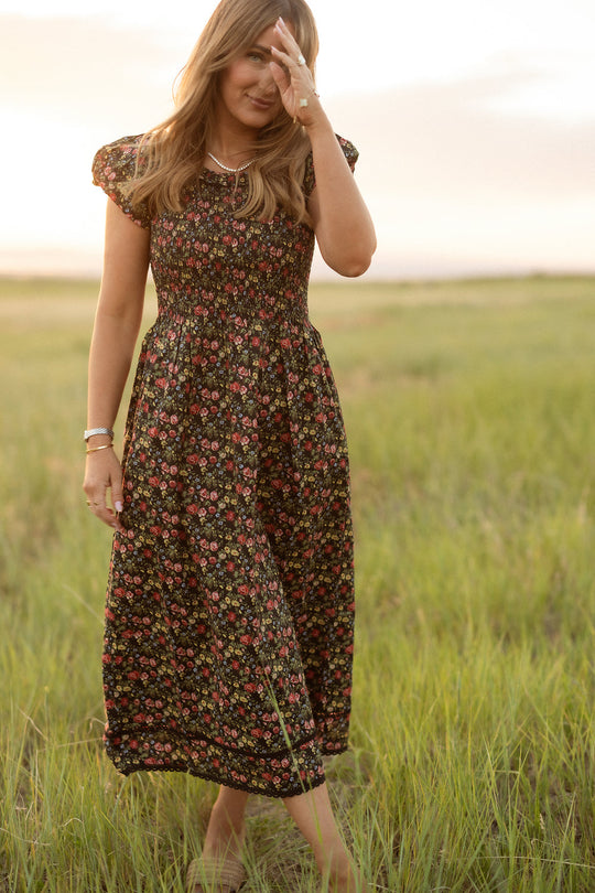 A woman wearing the Annalise Dress in Obsidian Vintage Poppies by Ivy City Co stands in a grassy field at sunset, one hand partially shielding her face while the other rests by her side. The maternity-friendly smocked bodice dress flows gracefully around her, perfectly capturing the dreamy mood of golden hour.