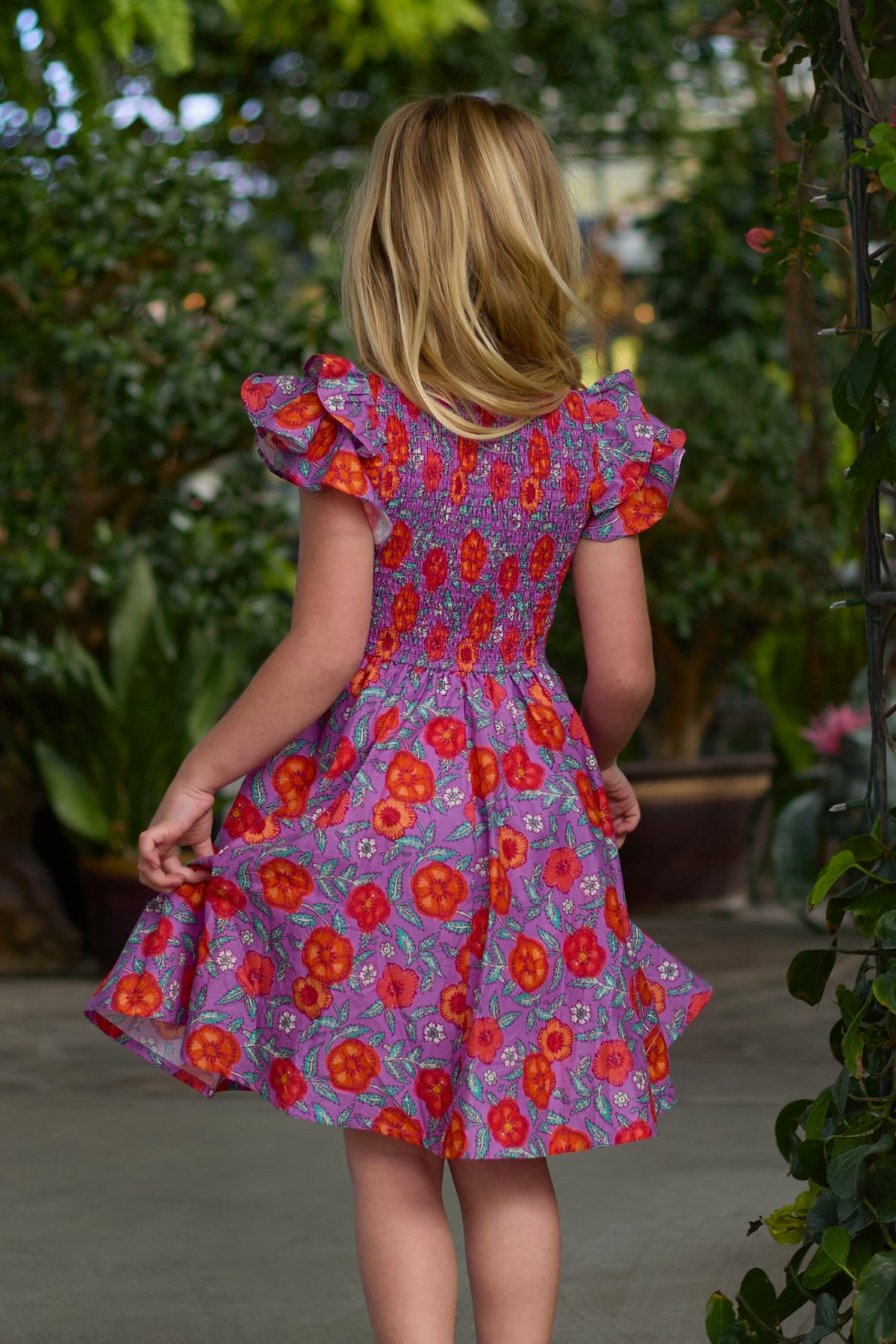 A young girl with blonde hair stands outdoors, surrounded by lush green plants, wearing the Ivy City Co Mini Hattie Dress in Thistle Flower Poppy Woodblock. The beautiful cotton midi dress features ruffled sleeves and a vibrant purple floral pattern with red and orange accents, highlighting its enchanting charm.
