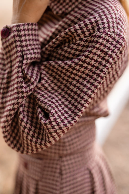 A close-up of a person wearing the Ivy City Co Bradley Jacket in Mauve Houndstooth, featuring ruffled sleeves and a bold purple and pink houndstooth pattern, with their arm bent toward their head. This nursing-friendly jacket beautifully blends eye-catching style with practical functionality.