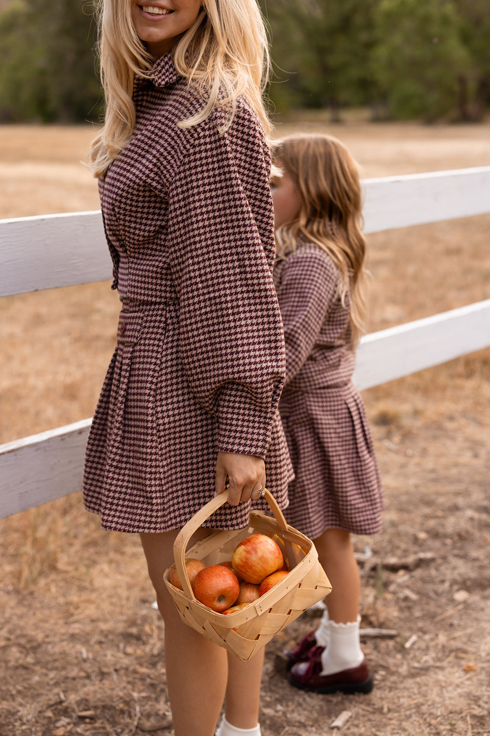A woman wearing the Ivy City Co Bradley Jacket in Mauve Houndstooth, which is nursing friendly, stands by a wooden fence in a field alongside a young girl. Both are dressed in matching plaid outfits; the woman holds a basket of apples while the girl stands beside her, facing away.