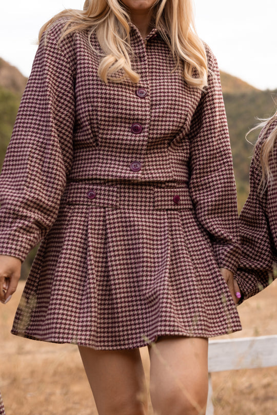 A person stands outdoors wearing the Ivy City Co Bradley Jacket in Mauve Houndstooth with a matching pleated skirt, their long blonde hair partially covering their face.