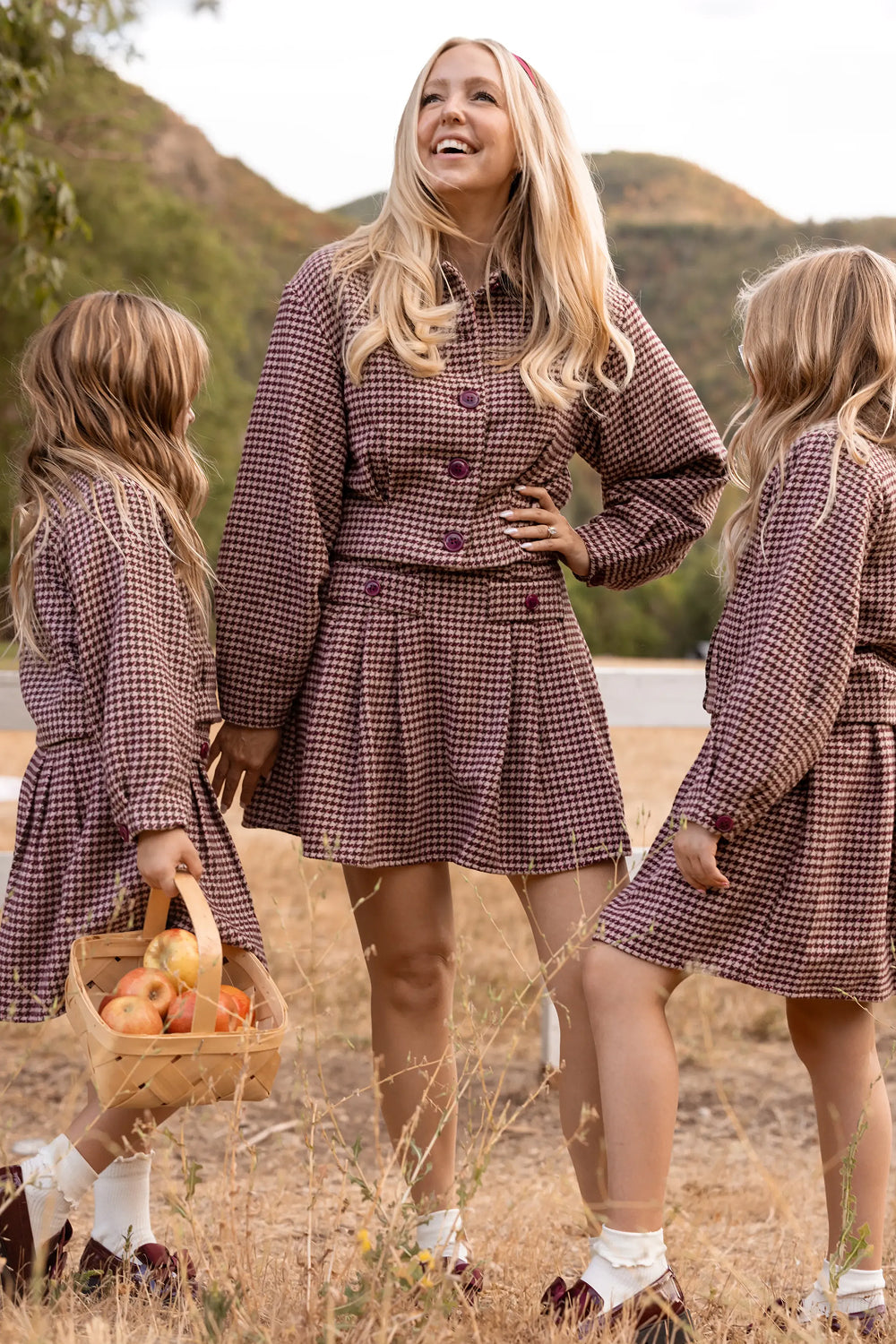 A woman wearing the Ivy City Co Bradley Jacket in Mauve Houndstooth stands outdoors with two young girls dressed in matching plaid outfits. Smiling, she holds hands with one girl while the other carries a basket of apples, with dry grass and hills visible in the background.