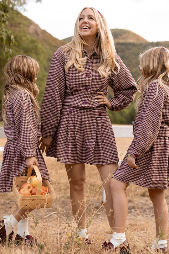 A woman wearing the Ivy City Co Bradley Jacket in Mauve Houndstooth stands outdoors with two young girls dressed in matching plaid outfits. Smiling, she holds hands with one girl while the other carries a basket of apples, with dry grass and hills visible in the background.