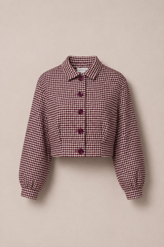 The Bradley Jacket in Mauve Houndstooth by Ivy City Co features a cropped silhouette with a maroon and white houndstooth pattern, purple buttons, a classic collar, and long, subtly puffed sleeves. This stylish Ivy City Co jacket is showcased against a neutral beige background.