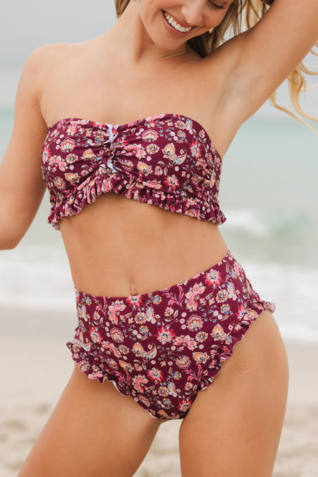A woman smiles on the beach in Ivy City Co’s High Waist Ruffle Bottom in Stephanie Floral, a maroon strapless bikini with floral pattern, ruffle details, and high-waisted bottoms, with ocean and sand in the background.
