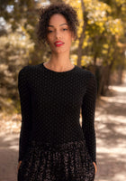 Woman wearing a black long-sleeve top with a pattern, standing outdoors with trees in the background.