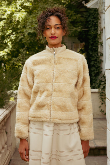Woman wearing a beige fur jacket outdoors with greenery in the background