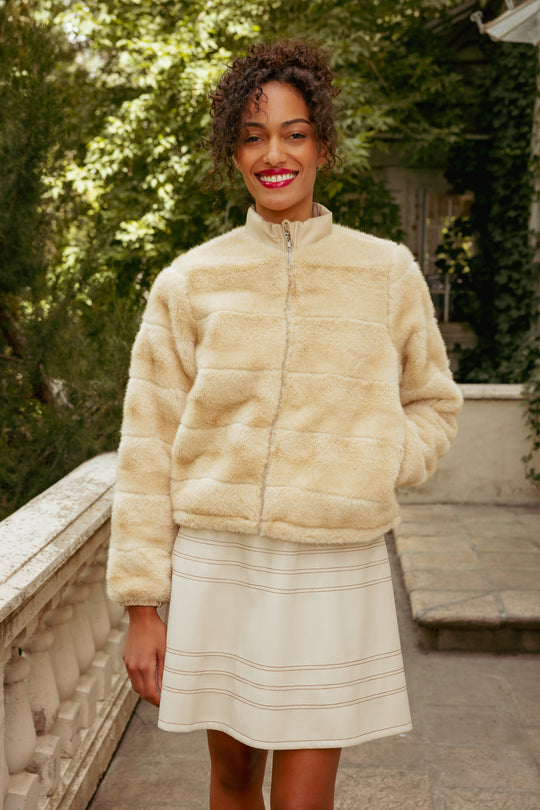 Woman wearing a beige fur coat and skirt standing outdoors with greenery in the background