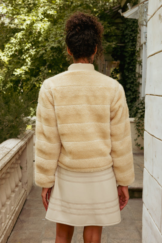 Person wearing a beige fur coat and skirt standing on a stone path with greenery in the background
