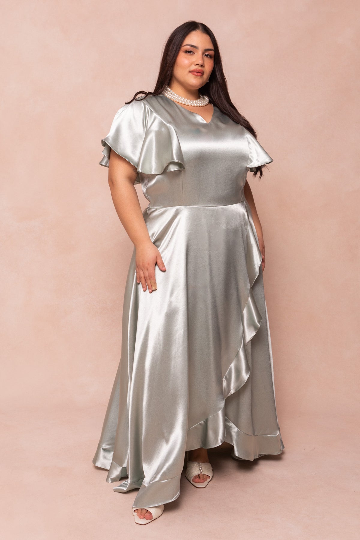 A woman models the Ivy City Co Callie Dress in Sage Satin, featuring short flutter sleeves and a ruffled skirt, paired with white open-toe heels and a pearl necklace against a soft beige background.