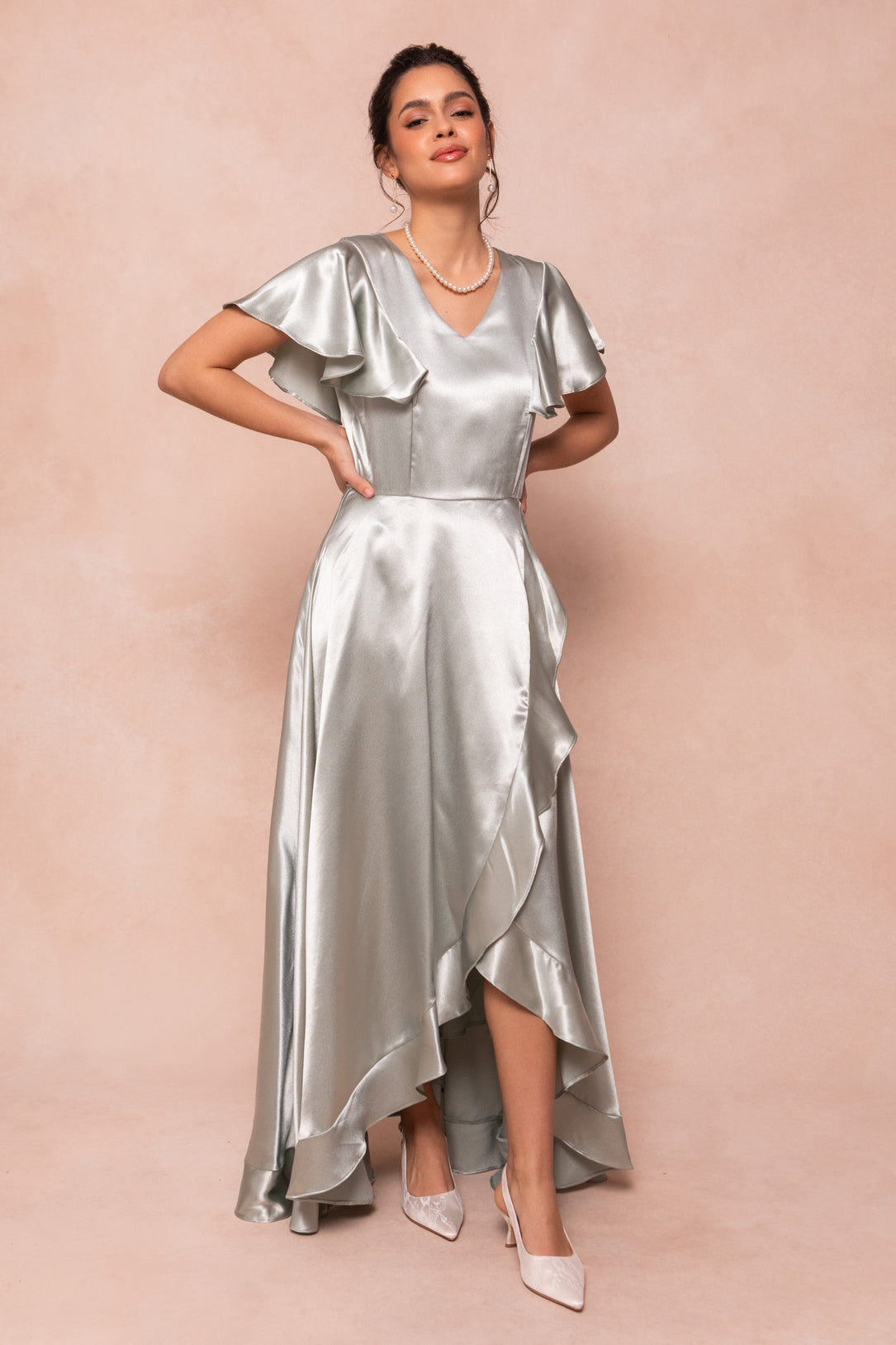 A woman wearing the Ivy City Co Callie Dress in Sage Satin, featuring ruffled short sleeves and an asymmetrical hem, stands against a light peach background, smiling with her hands on her hips. She pairs it with light heels and a necklace.