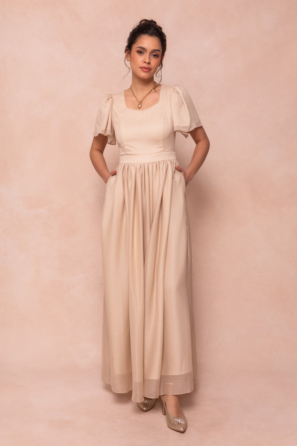 A woman stands against a soft pink background, wearing the Ivy City Co Abbie Dress in Champagne. This maxi dress features short puff sleeves, a square neckline, a fitted waist, and pockets. She completes the bridal look with gold shoes and a necklace.