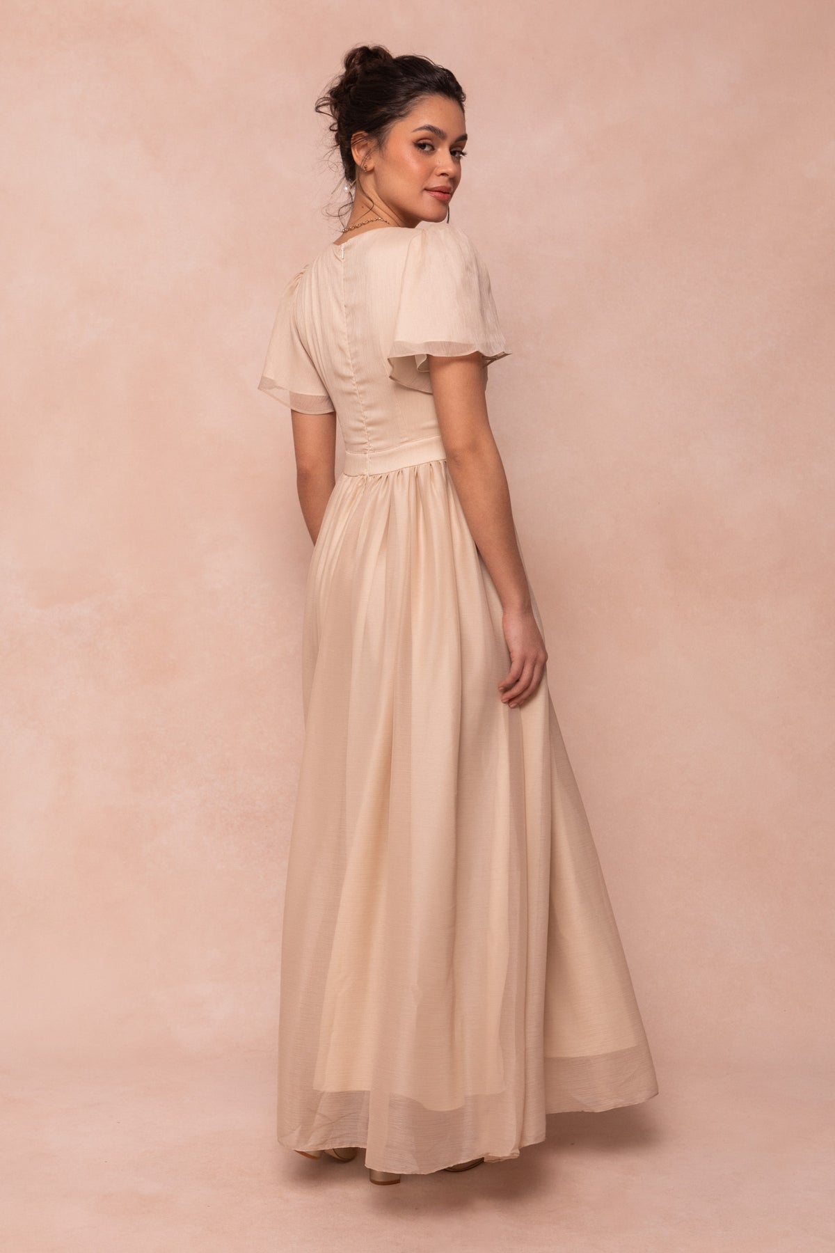 A woman with dark hair in an updo wears the Ivy City Co Abbie Dress in Champagne—a maxi with short puff sleeves—turned to the side and looking over her shoulder against a soft beige background, perfect for bridal parties.