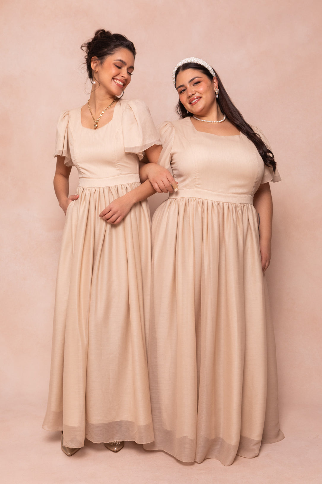 Two women stand side by side in Ivy City Co's Abbie Dress in Champagne—maxi dresses with puff sleeves and square necklines—smiling and linking arms, accessorized with necklaces and earrings, against a soft light pink backdrop. Perfect for bridal parties.