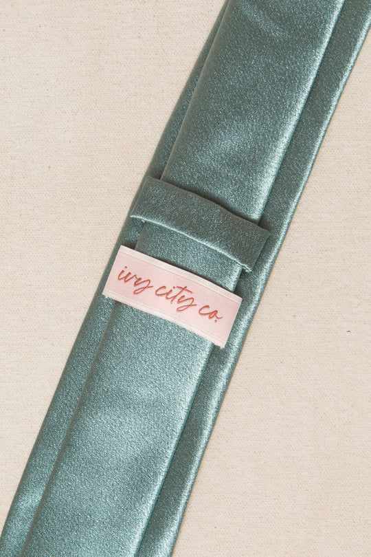 A close-up of the Mens Max Tie in Seaglass Satin by Ivy City Co, featuring metallic green satin fabric and a pink tag with the brand's red script, displayed on a light beige background.
