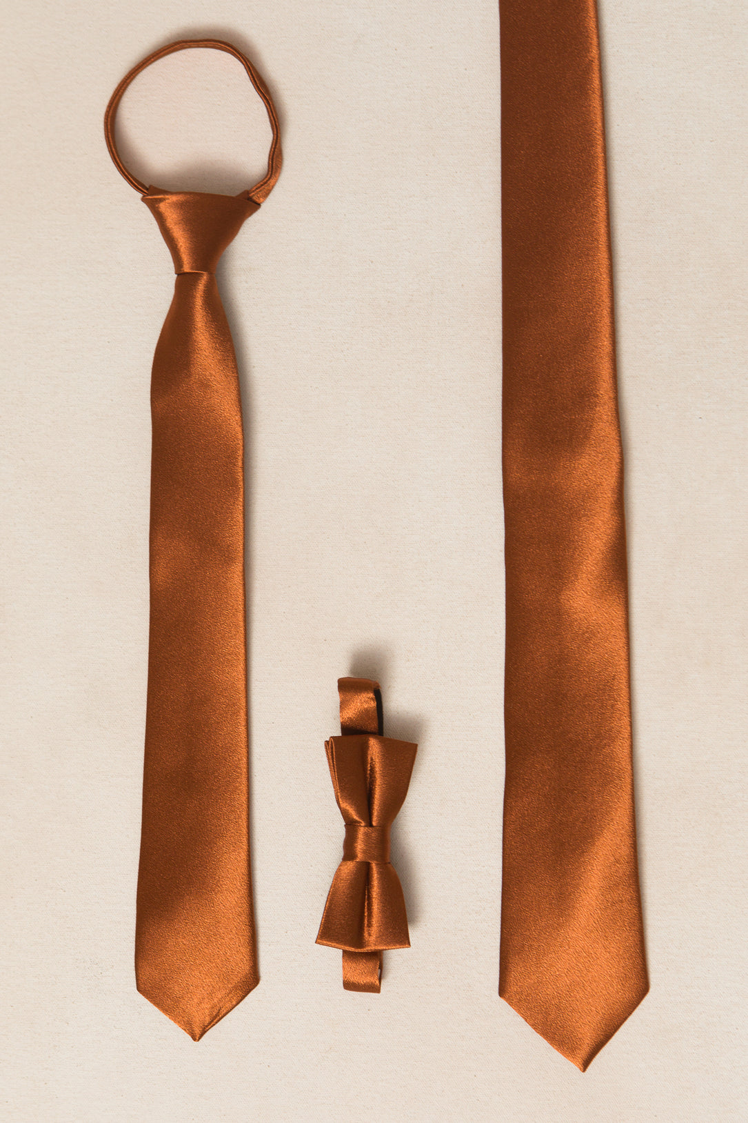 Two brown satin neckties—a Mens Max Tie in Amber Satin by Ivy City Co and a self-tie—are placed vertically on a beige surface. A matching brown satin bow tie lies horizontally between them.