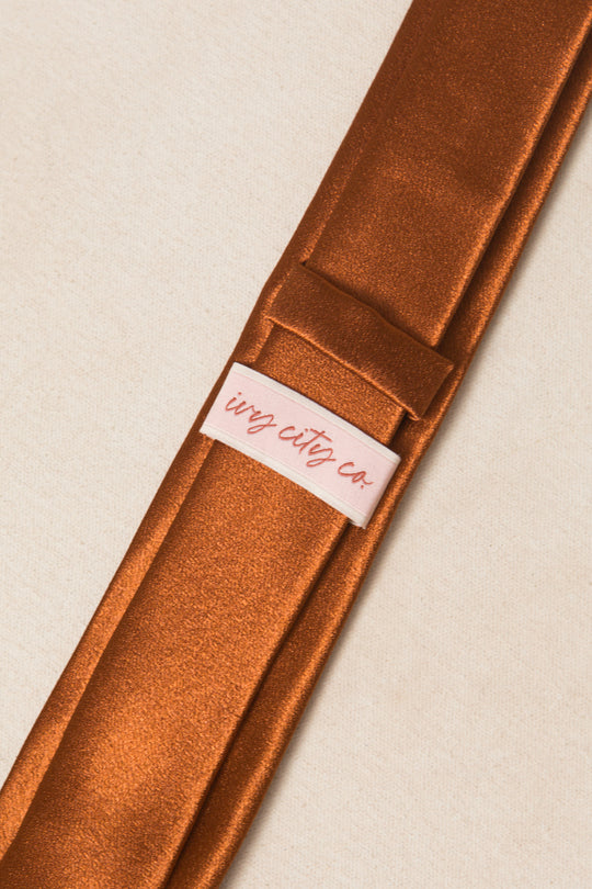 A close-up of the Mens Max Tie in Amber Satin by Ivy City Co, featuring a shiny brown satin fabric and a pink Ivy City Co label, set against a cream-colored background.