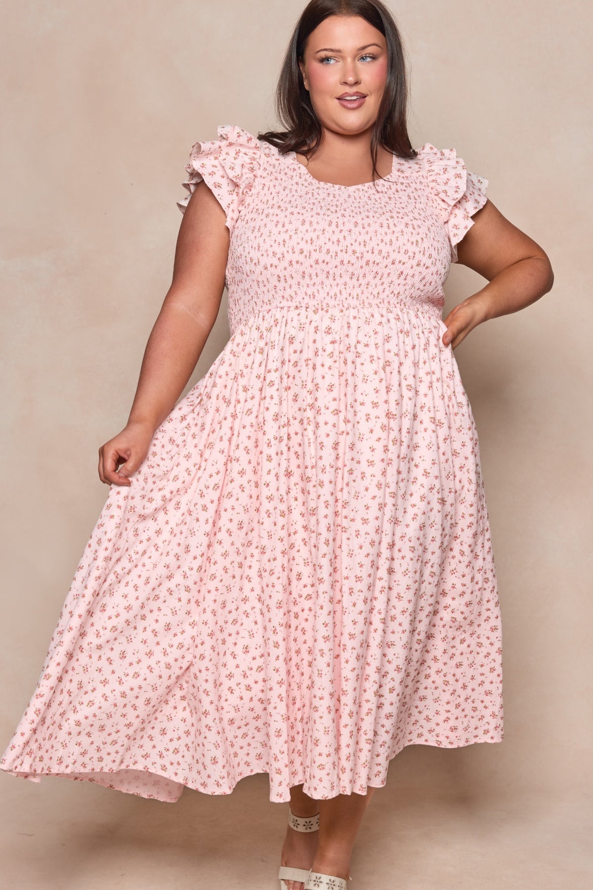 A woman wearing the Hattie Dress in Madeline Pink by Ivy City Co—a light pink, floral-print cotton midi dress with ruffled sleeves—stands against a neutral background, holding out the skirt and smiling.